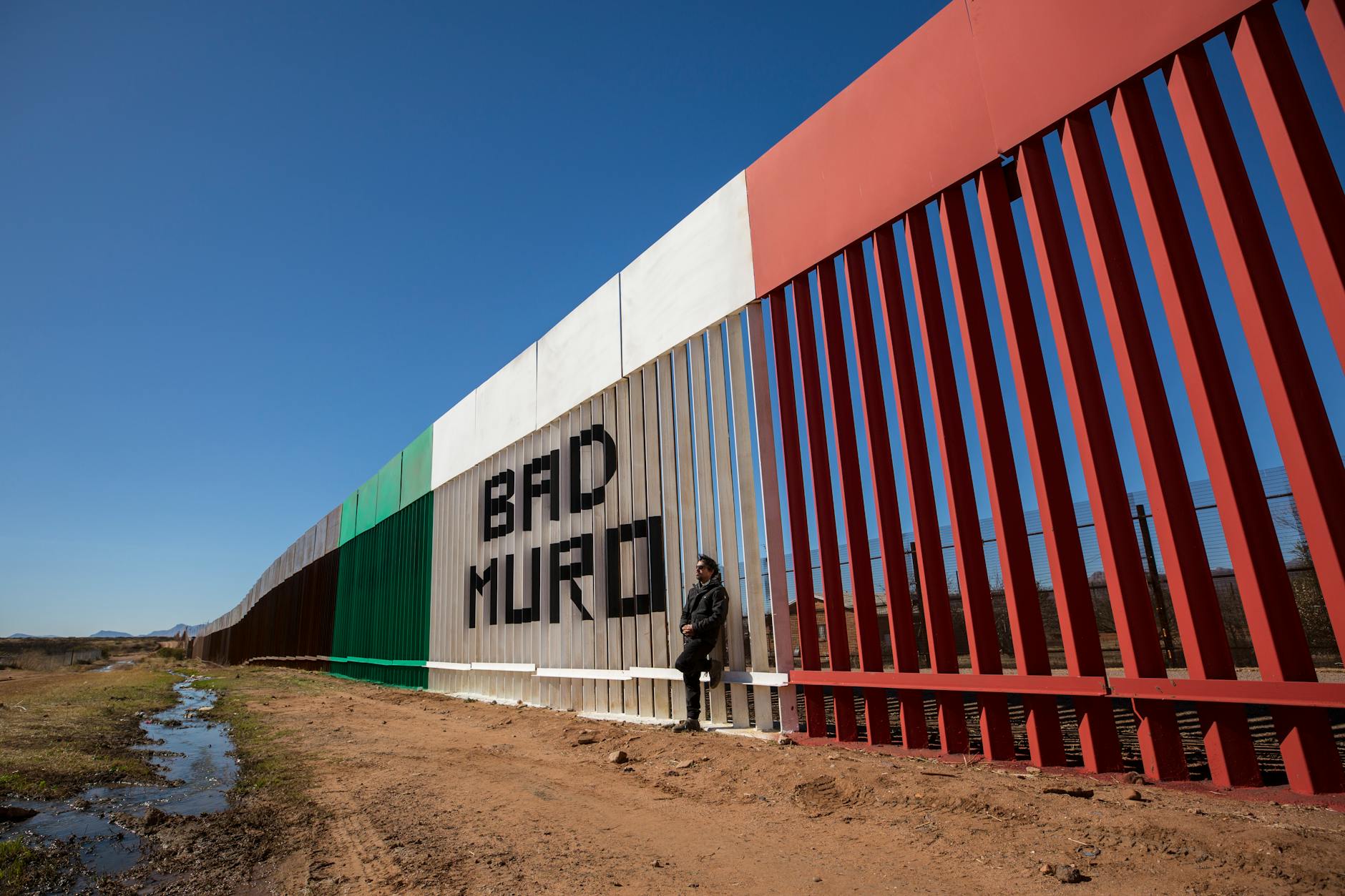 Image of a man standing by the border wall painted with Mexican flag colors in Naco, Sonora, Mexico.